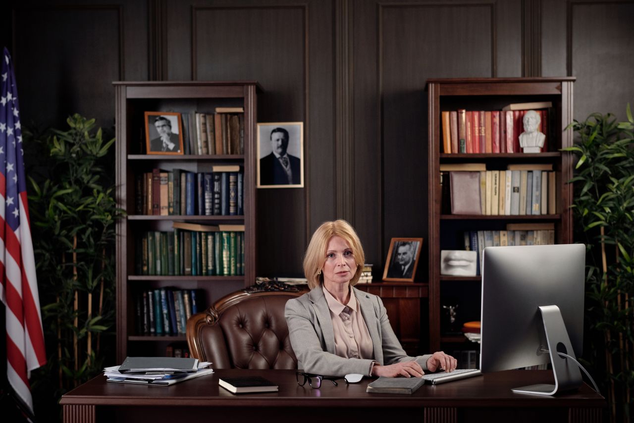 The Art of Drawing Readers In: Your attractive post title goes here A businesswoman sits confidently at her desk in an office setting with a bookshelf and American flag.